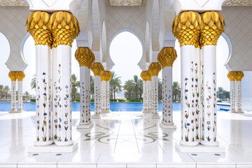 A white marbe colonnade with golden capitals leading to the external pool with palm trees on the background at Sheik Zayed mosque