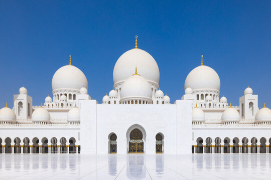Symmetrical close up of the main courtyard of the Sheik Zayed grand mosque in Abu Dhabi with the prayer hall and its round domes against a blue sky