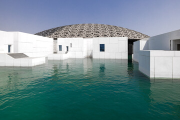 Exterior of the modern building of Louvre in Abu Dhabi, with the latticework dome and the white marble wall, surrounded by water