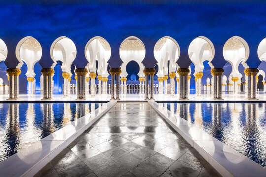 Symmetrical nightshot of the colonnade of the Sheik Zhayed mosque, with a marble catwalk surrounded by water