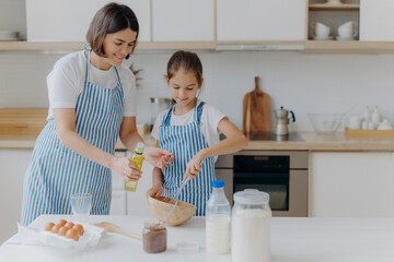 Brunette mother adds oil in dough, little daughter helps to make pastry, whisks ingredients, pose together against kitchen interior, prepare bakery together. Small helper with mommy. Homemade food