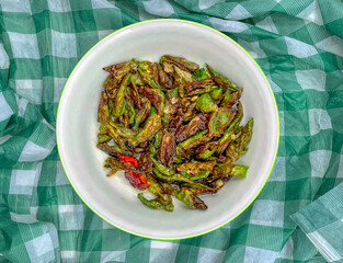 Fried green peppers and one red with olive oil in a bowl on a green and white checkered cloth tablecloth