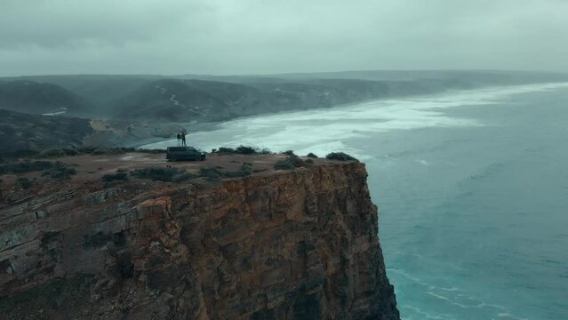 Aerial drone shot of couple stand on top of adventure van at edge of cliff on epic and dramatic coastline. Cinematic shot of moody travel destination. Vanlife lifestyle for millennials