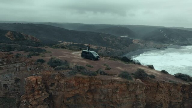 Aerial drone shot of couple stand on top of camper van, hold hands and look out into distance. Inspiring vanlife offroad adventure. Urban nomads living offgrid in overlanding vehicle
