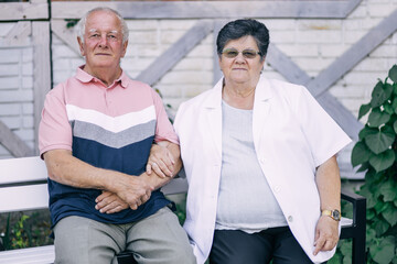 Grandfather and grandmother are sitting on a bench near the house. An elderly woman with glasses holds a man's hand