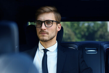 Thoughtful confident businessman in full suit with eyewear looking away while sitting in the car.