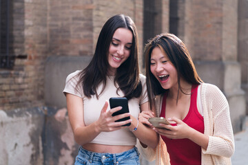 two young women laughing funny looking at their cell phones as they stroll through the city arm in...