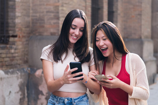 two young women laughing funny looking at their cell phones as they stroll through the city arm in arm during a date, concept of friendship and love between people of the same sex