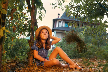 Country summer. Dreaming caucasian little girl wearing straw hat sits on the ground in garden and looking at the sky. The concept of psychology