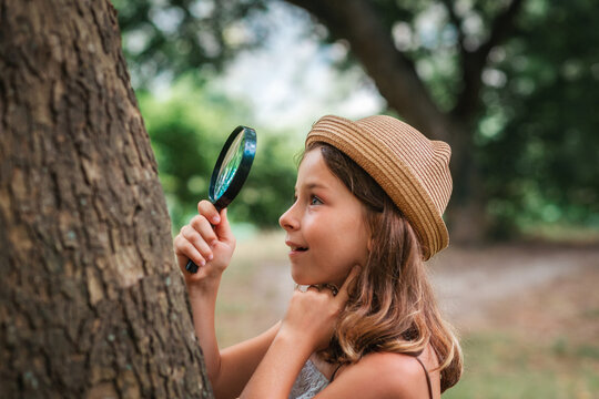 Children's education. Portrait of amazement little girl in a straw hat looks at the tree bark through a magnifying glass. The concept of scouting and curious childhood