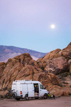ALABAMA HILLS, LONE PINE, CA, USA. A white van parked against large rounded boulders at a secret camp spot with moon rising in lavender sky.
