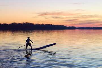 Fototapeta na wymiar Silhouette of young man standing on SUP (stand up paddle board) at autumn sunset in the Danube river. Water sport at blue hour