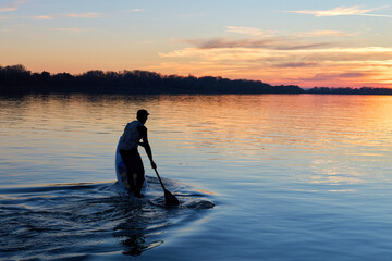 Silhouette of young man standing on SUP (stand up paddle board) at autumn sunset in the Danube river. Water sport at blue hour