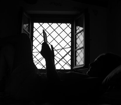 black and white woman silhouette resting lying on a bed while holds an electronic book with her hands and looks at the screen to read the book. At the background light comes throug