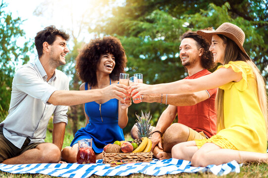 Group of friends having pic-nic in a park on a sunny day - Happy young men and women toasting healthy orange fruit juice at springtime camping - People having fun and relaxing in summer holidays.