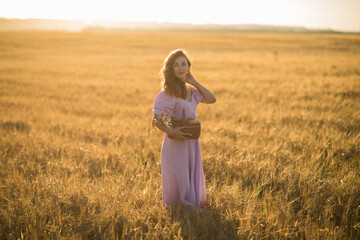 a girl with long dark hair in a lilac dress with a basket in her hands at sunset in a wheat field