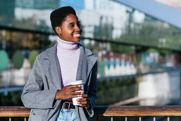 Cheerful black female in stylish outerwear with zero waste cup of coffee smiling and looking away....