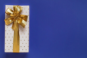 White gift box with golden ribbon and bow on a blue background. Top view. Copy space.