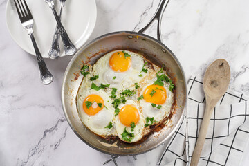 Four fried eggs with unbroken egg yolks, fresh green herbs on a metal dirty pan on a marble background, top view