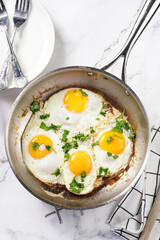 Four fried eggs with unbroken egg yolks, fresh green herbs on a metal dirty pan on a marble background, top view