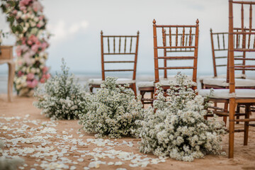 Wedding wooden chairs decorated with flowers. Rustic aisle chairs standing on sand for ceremony on the beach. Natural, shabby, boho wedding decor