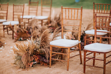 Wedding wooden chairs decorated with flowers. Rustic aisle chairs standing on sand for ceremony on the beach. Natural, shabby, boho wedding decor - 561507953
