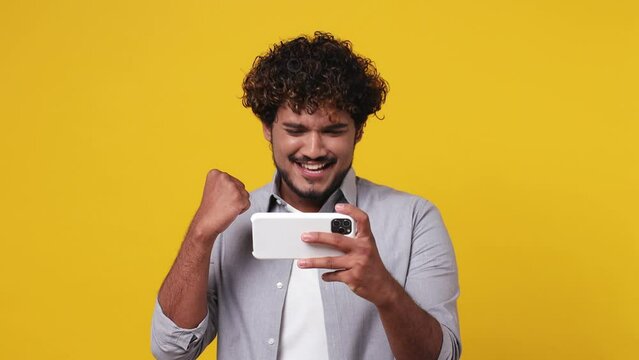 Gambling excitable young Indian man in casual grey shirt use play racing app on mobile cell phone hold gadget smartphone for pc video games do winer gesture isolated on plain yellow background studio