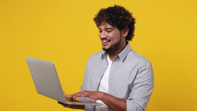 Smart IT user smiling happy cheerful young Indian man 20s wear casual grey shirt hold in hand use work typing on laptop pc computer isolated on plain yellow background studio. People lifestyle concept