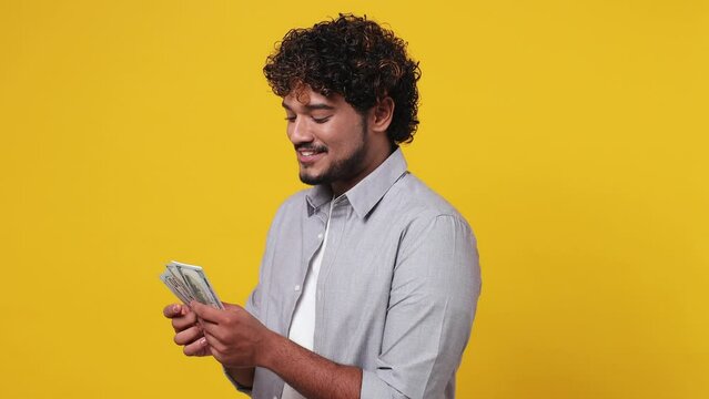 Sideways accountant satisfied young Indian man wear casual grey shirt hold in hand count stack of cash money in dollar banknotes isolated on plain yellow background studio. People lifestyle concept