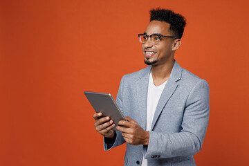 Young employee business man corporate lawyer wears classic formal grey suit shirt glasses work in office use digital tablet pc computer look aside area isolated on plain red orange background studio.