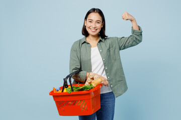 Young happy woman wears casual clothes hold red basket with food products show hand biceps muscles demonstrate power isolated on plain blue background studio. Delivery service from shop or restaurant.