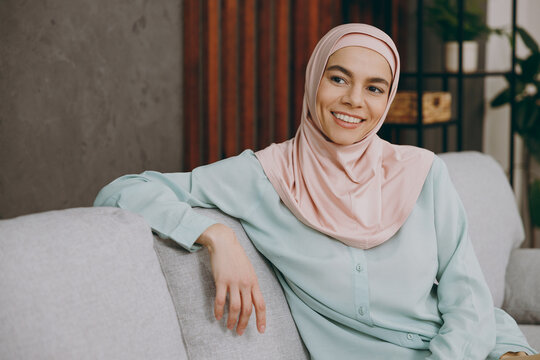 Young minded calm muslim woman wear hijab casual clothes look aside sits on sofa couch stay at home flat rest relax spend free spare time in living room indoor People uae middle eastern islam concept