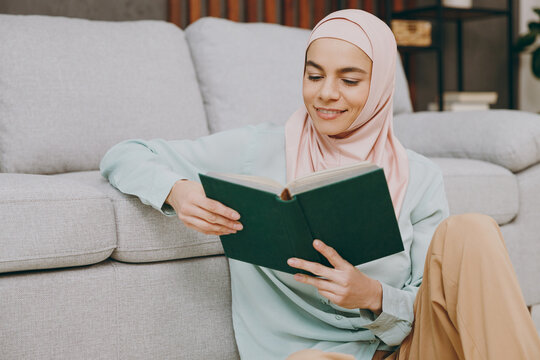 Young calm muslim woman wearing hijab casual clothes reading book sit near sofa couch stay at home flat rest relax spend free spare time in living room indoor. People middle eastern uae islam concept.