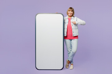 Full body elderly blonde woman 50s year old wear casual clothes denim jacket t-shirt point finger on big huge blank screen mobile cell phone with area isolated on plain pastel light purple background