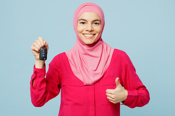 Young smilng arabian muslim woman wears pink abaya hijab hold car keys fob keyless system show thumb up isolated on plain pastel light blue cyan background studio People uae islam religious concept.