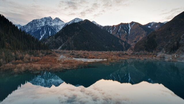 Issyk mountain lake with mirror water at sunset. The color of the water changes before our eyes. There are trees in clear water. Snowy mountains and green hills are visible. Clouds are reflected © SergeyPanikhin