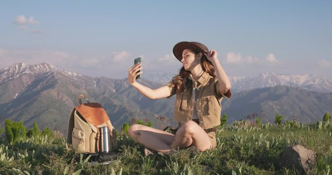 Woman hiker takes a selfie on smartphone. Millennial woman hiker adjusts her hat while taking selfie against the background of picturesque mountain landscape to get interesting and beautiful photos. 