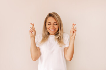 A young attractive smiling blonde woman in a white t-shirt with her eyes closed crosses her fingers for good luck waiting for the results of the lottery or exams isolated on a color beige background