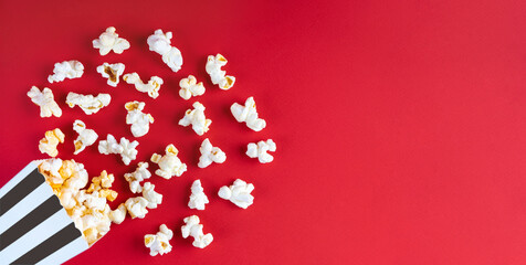 Tasty cheese popcorn falling out of a black striped carton bucket, isolated on red background. Scattering of popcorn grains. Movies, cinema, fast food and entertainment concept. Top view, flat lay