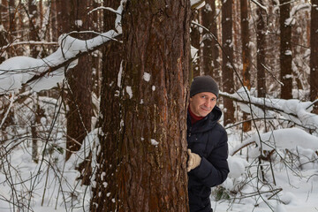 Portrait of young millennial smiling man peeking out from pine trunk in winter coniferous forest.