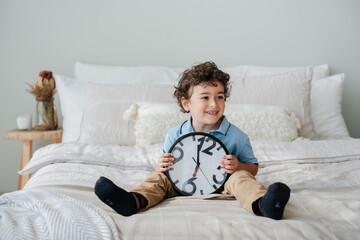 Handsome  curly Spanish little boy in blue pole holds wall clock smiles sits on bed. Playful baby boy happy at home learns numbers, waits for parents. Cute children. Childhood, domestic family leisure