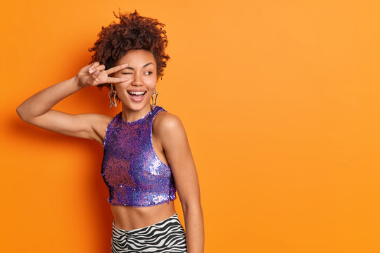 Horizontal shot of positive curly haired woman shows peace sign smiles broadly winks eye wears purple top has slim figure isolated over orange background blank space for your promotional content