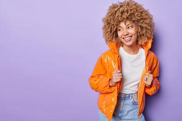 Indoor shot of positive thoughtful woman with curly hair smiles broadly looks aside dressed in...