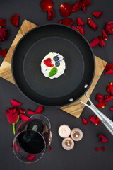 Valentine's day concept with red rose flower petals on a black background and a pan placed on a wooden board and a pavlova cake inside the pan. A glass of red wine next to it. View from above.