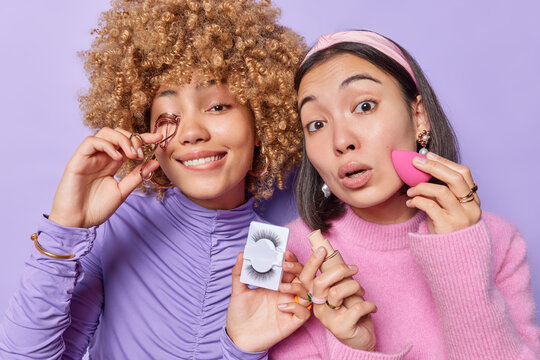 Horizontal shot of two female models apply foundation on face holds eyelashes put on makeup prepare for party dressed casually isolated over purple background. Women and beauty treatments concept