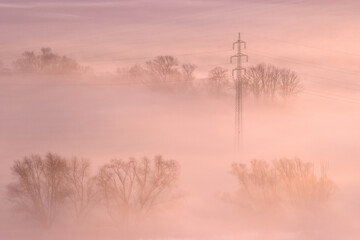 High voltage power lines emerging from ground fog in an agricultural landscape. Pastel colours of sunrise, warm tones, ground fog, frosty morning. Czech Republic.