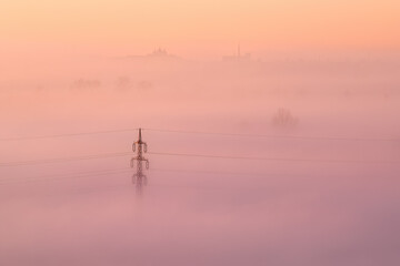 High voltage power lines emerging from ground fog in an agricultural landscape. Pastel colours of...