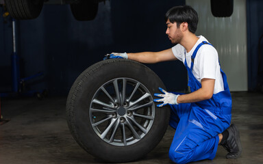 Fototapeta na wymiar Asian male mechanic wearing uniform, changing rubber tyre or tire wheel, working in garage at car or automobile maintenance service center or shop. Industry Concept.