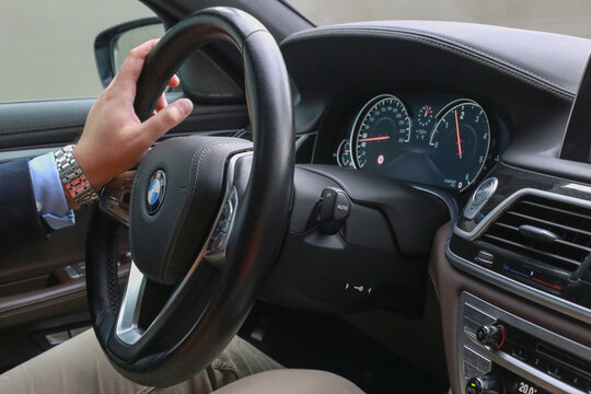 BONN, GERMANY - November 2021: BMW 7 Series central console panel, car steering wheel close-up. Man is driving car against gray wall, background. The businessmans hand holds the steering wheel.