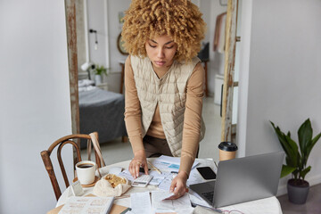 Indoor shot of curly haired woman stands near table full with papers invoices cheque bills does paperwork calculates expenses uses calculator wears jumper and vest manages monthly household incomes